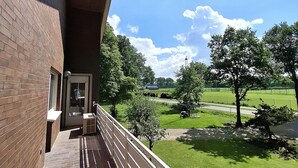 A balcony with a brick wall, a metal railing, and a view of a green park with trees and a clear sky. - "Anno 1973" - Start your journey through time today! PRIMA Quarter (Lastrup)