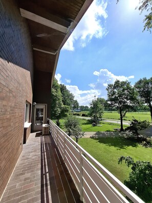 Ein Balkon mit Backsteinmauer, Metallgeländer und Blick auf einen grünen Park mit Bäumen und blauem Himmel.