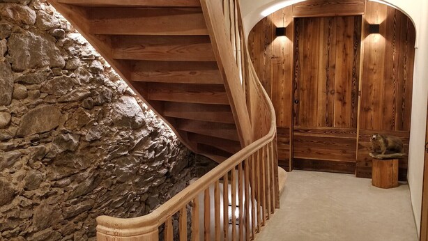 Una scala in legno con un muro in pietra su un lato, che conduce a una stanza con pannelli in legno e un piccolo sgabello di legno.