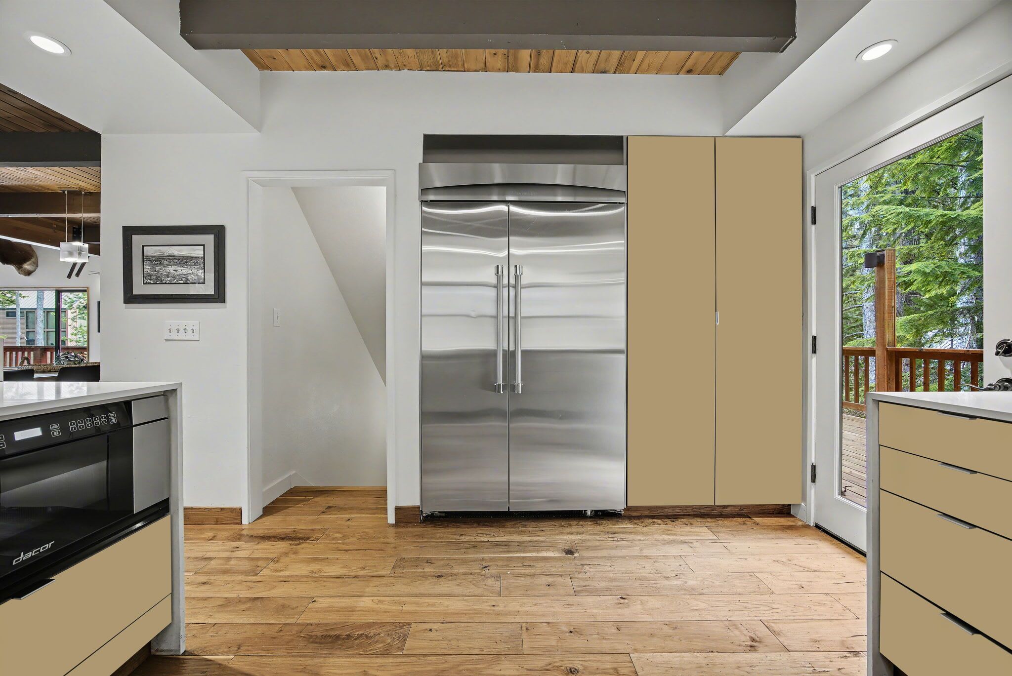Eine moderne Küche mit einem Edelstahl-Kühlschrank, Holzboden und Blick auf eine Terrasse durch eine Glastür.