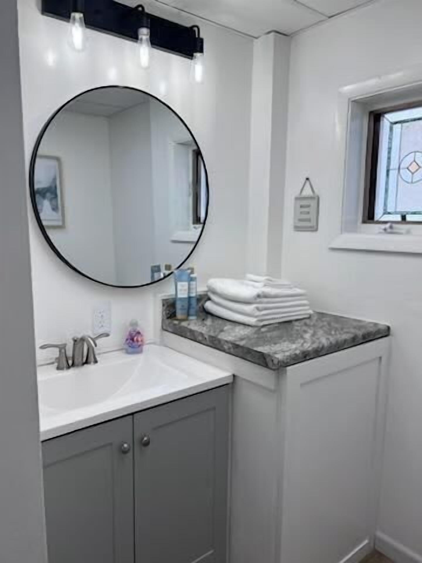Une salle de bain avec un miroir rond, un lavabo, une fenêtre et un porte-serviettes.