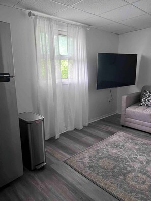 A room with a television mounted on the wall, a sofa, a trash bin, and a window with curtains.