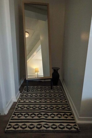 A hallway with a patterned rug, a tall mirror, a lamp, and a vase.