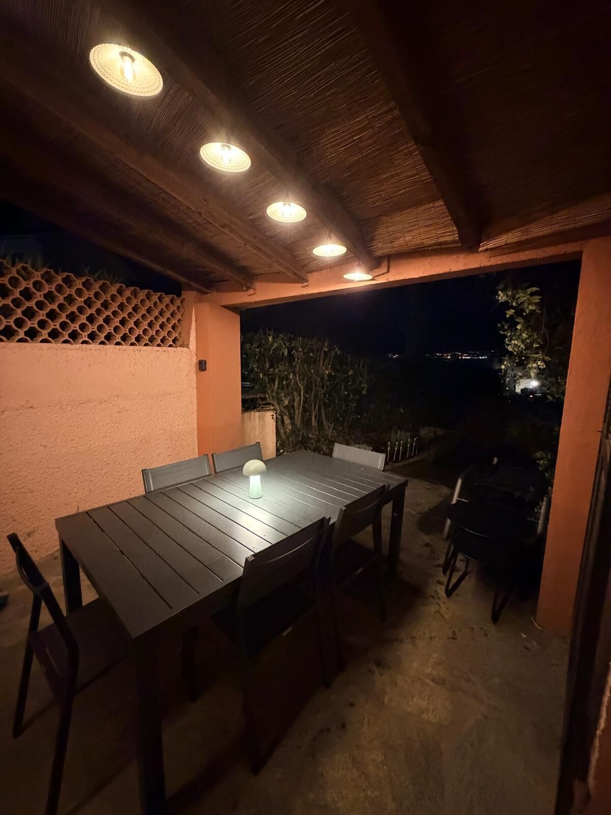 An outdoor patio with a wooden table and chairs, a hanging light fixture, and a view of the night sky.