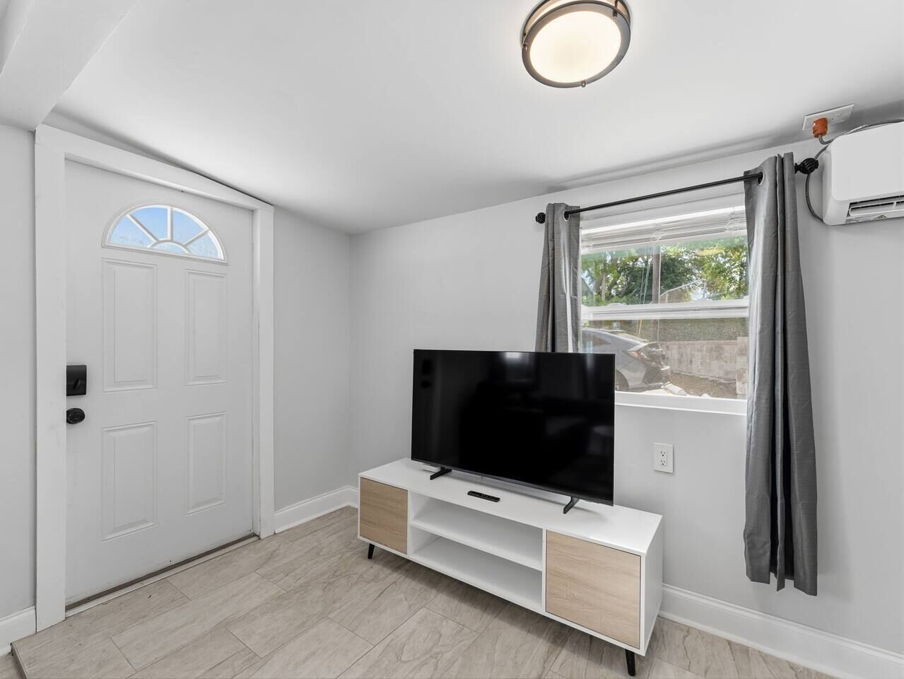 Une pièce avec une porte blanche, un téléviseur à écran plat posé sur un meuble télé blanc, une fenêtre avec des rideaux et un luminaire au plafond.