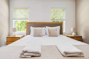 Ein ordentlich gemachtes Bett mit weißen Leinen und Kissen, zwei Nachttische mit Lampen sowie Fenster mit Jalousien.