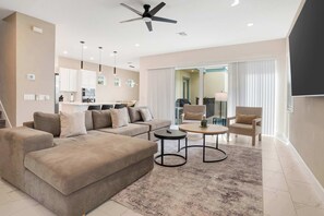 Ein modernes Wohnzimmer mit Sofa, Couchtisch und Sesseln, im Hintergrund eine Küche und ein Deckenventilator.