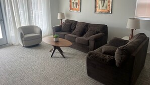 A living room with a sofa, armchair, and a small table with a plant. There are two wall-mounted lamps and two framed pictures. - OCEANFRONT PENTHOUSE (Wildwood Crest)