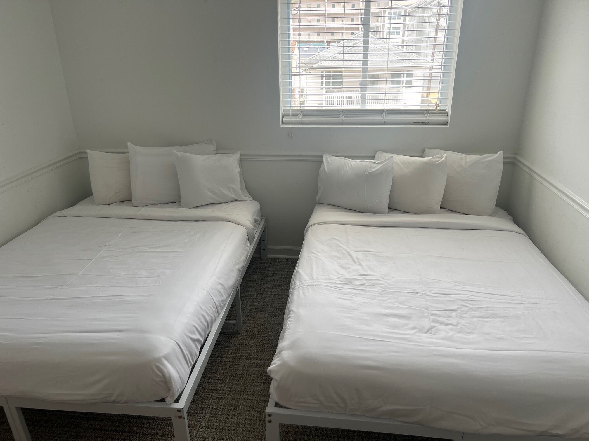 Ein Doppelzimmer mit zwei Einzelbetten, einem Fenster mit Jalousien und einer schlichten Wand.