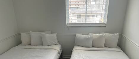 Ein Doppelzimmer mit zwei Einzelbetten, einem Fenster mit Jalousien und einer schlichten Wand.