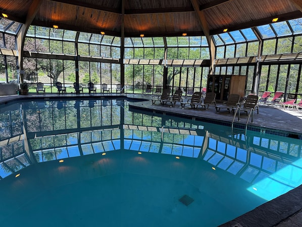 Ein großer, überdachter Pool mit Glaswänden, umgeben von Liegestühlen und einer Holzbalkendecke.