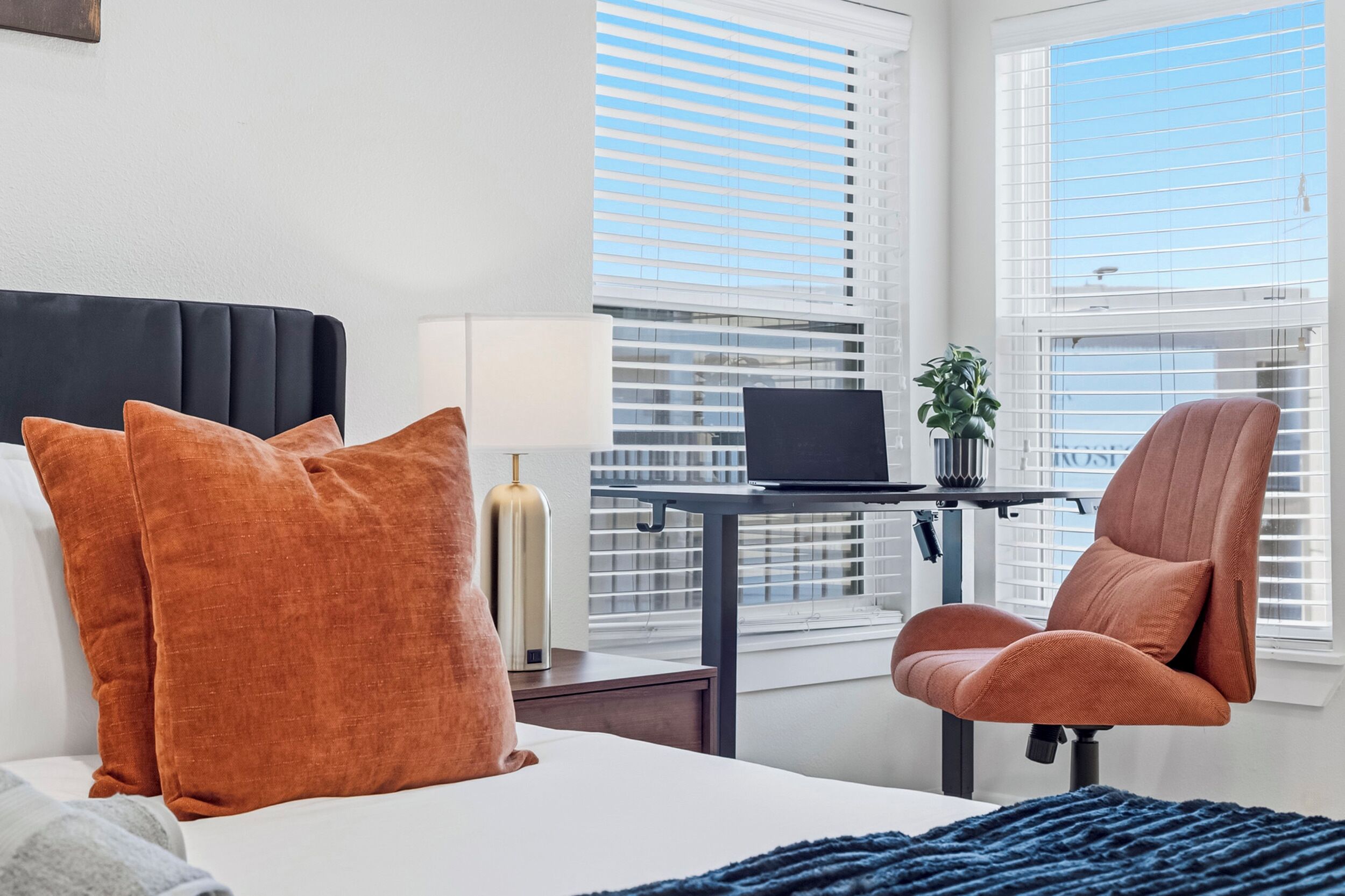 Une chambre d’hôtel avec un lit, un bureau avec un ordinateur portable, une chaise et une fenêtre avec des stores.