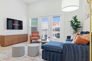 A modern living room with a flat-screen TV, a sofa, ottomans, a coffee table, and a potted plant. - Uptown 2BR Shops, Dining, Katy Trail, Parking (Dallas)