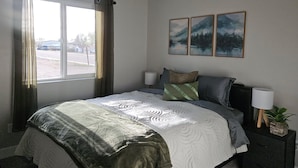 Habitación de hotel con una cama, ventilador de techo, ventana con cortinas, mesita de noche con lámpara y tres cuadros en la pared.
