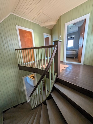 Una escalera con barandilla de madera que lleva a una plataforma, un pequeño cofre de almacenamiento, y un dormitorio con ventana y puerta.