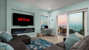 Ein Wohnzimmer mit Fernseher, der 'NETFLIX' anzeigt, Kamin, Schreibtisch mit Computer, Balkon mit Aussicht und großem Fenster.
