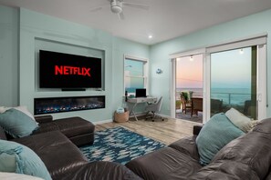 Ein Wohnzimmer mit Fernseher, der 'NETFLIX' anzeigt, Kamin, Schreibtisch mit Computer, Balkon mit Aussicht und großem Fenster.
