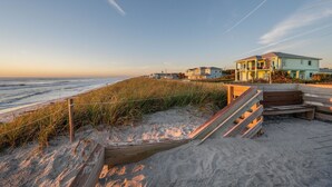 Ein Strandgrundstück mit einer Holztreppe, die zu einem Sandbereich führt, einem Holzzaun und einem hellblauen Haus mit einem Balkon.