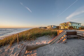 Ein Strandgrundstück mit einer Holztreppe, die zu einem Sandbereich führt, einem Holzzaun und einem hellblauen Haus mit einem Balkon.