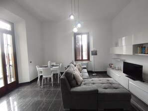 A modern living room with a grey sectional sofa, a dining table with chairs, a TV, and a bookshelf.