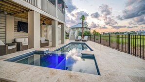 A swimming pool with a glass bottom, a covered patio area with outdoor furniture, and a view of a residential building in the background. - Private pool, putting green, kayaks, beautiful sunsets, ocean & marsh views! (Fripp Island)