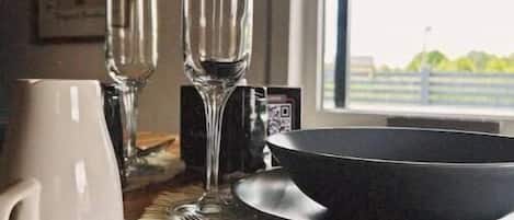 A dining table with a white mug, two wine glasses, a black bowl, and silverware on a wooden table with a woven placemat.