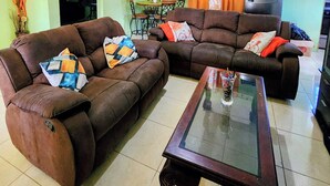 A living room with two brown sofas, a glass coffee table, a dining area with a table and chairs, and a TV on the wall.