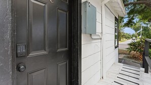 Una puerta negra con cerradura electrónica, un cuadro de empalmes eléctricos gris y una pared exterior blanca con una ventana.