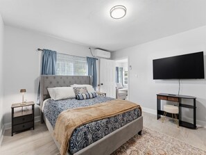 Ein Schlafzimmer mit einem Bett, Nachttischen, einem Schreibtisch, einem Stuhl, einem Fernseher und einem Fenster mit Vorhängen.
