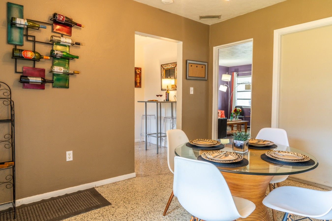 A dining area with a glass table, white chairs, and a wall-mounted wine rack with various bottles.