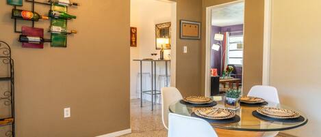 A dining area with a glass table, white chairs, and a wall-mounted wine rack with various bottles.