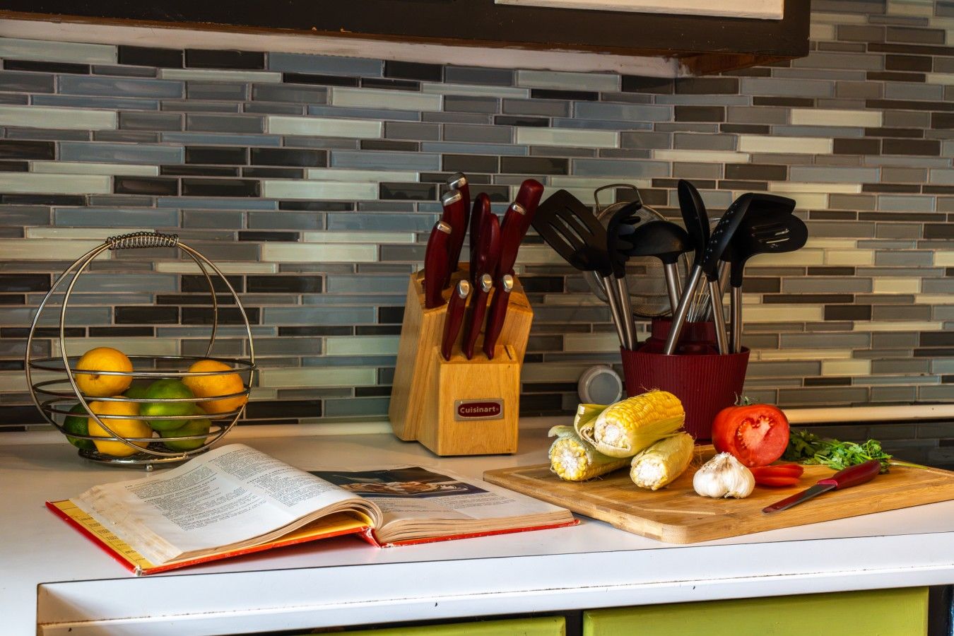 Un piano di lavoro in cucina con un libro, un cesto di frutta, un tagliere con verdure e utensili da cucina.