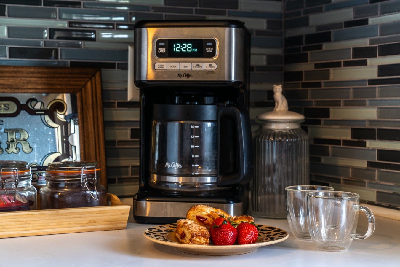 A coffee maker with a digital clock displaying 12:28 PM, a plate with pastries and strawberries, and a clear glass mug on a kitchen counter.