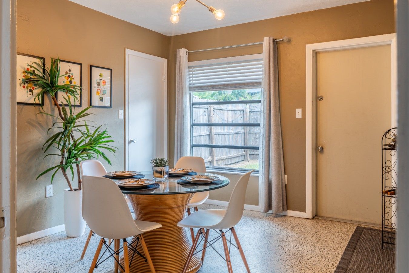 A dining area with a round table, four chairs, a potted plant, and framed artwork on the wall.