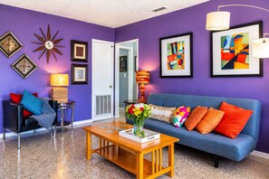 A living room with a blue sofa, a coffee table, and a door leading to another room. - Coastal Retro Haven: Private Yard • Near Beach (Sarasota)