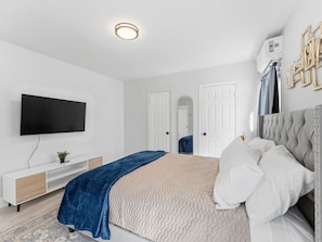 Ein modernes Schlafzimmer mit Flachbildfernseher, einem Bett mit weißer und grauer Bettwäsche, einer blauen Tagesdecke, einem weißen Konsolentisch und einem großen Spiegel.