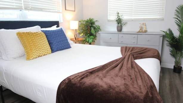 Een net opgemaakt bed met witte lakens, een bruine plaid en twee kussens met verschillende patronen.