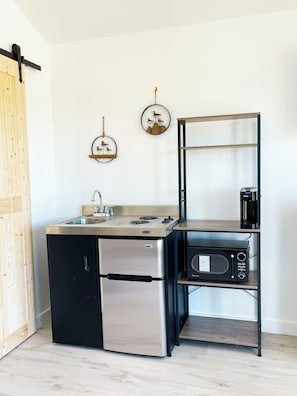 A compact kitchenette with a stainless steel refrigerator, a built-in stove, and a microwave on a wooden shelf.