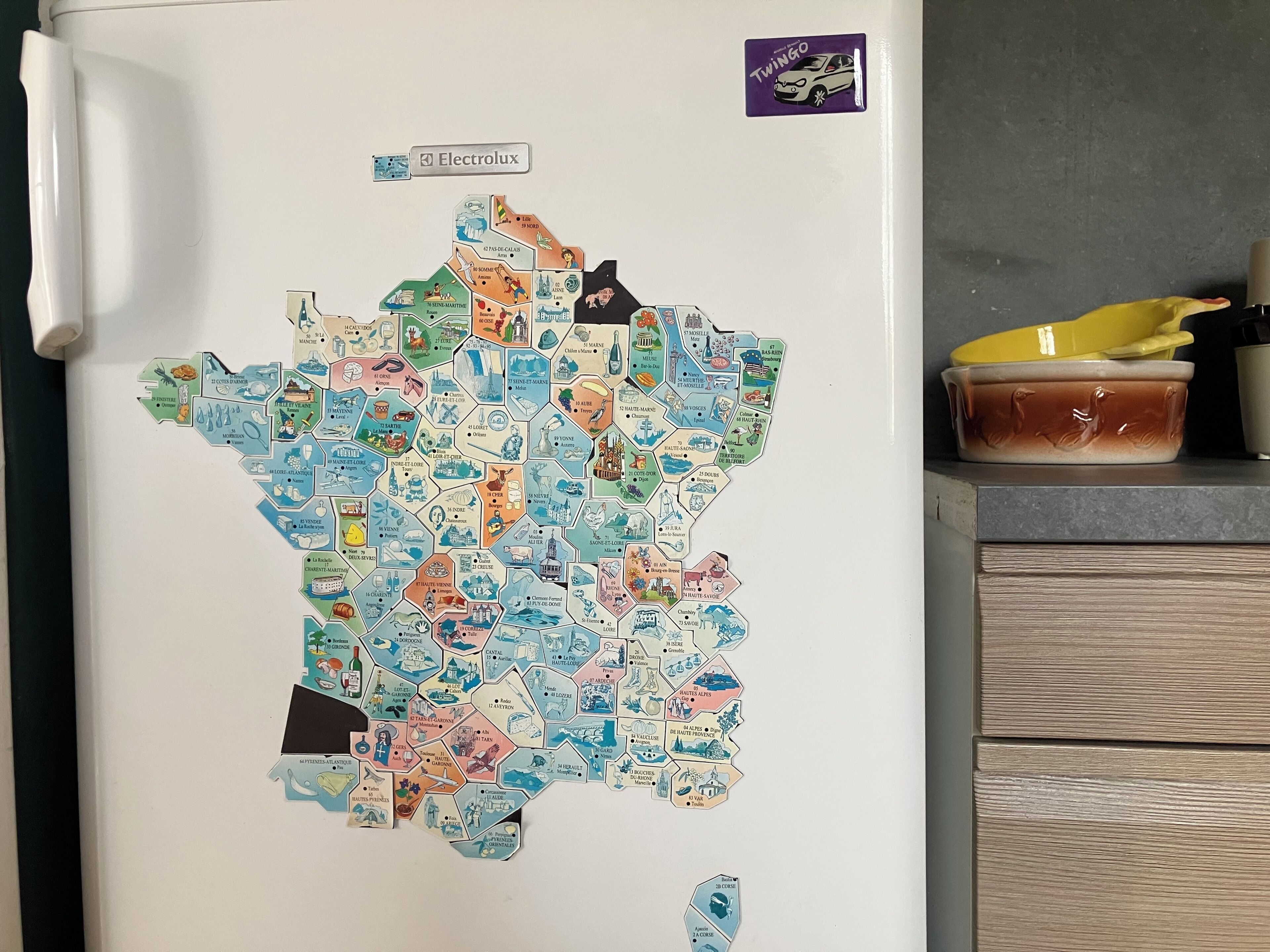 Un réfrigérateur dont la porte est ornée d’une carte de France, un autocollant représentant une Twingo et un récipient jaune posé sur le plan de travail.