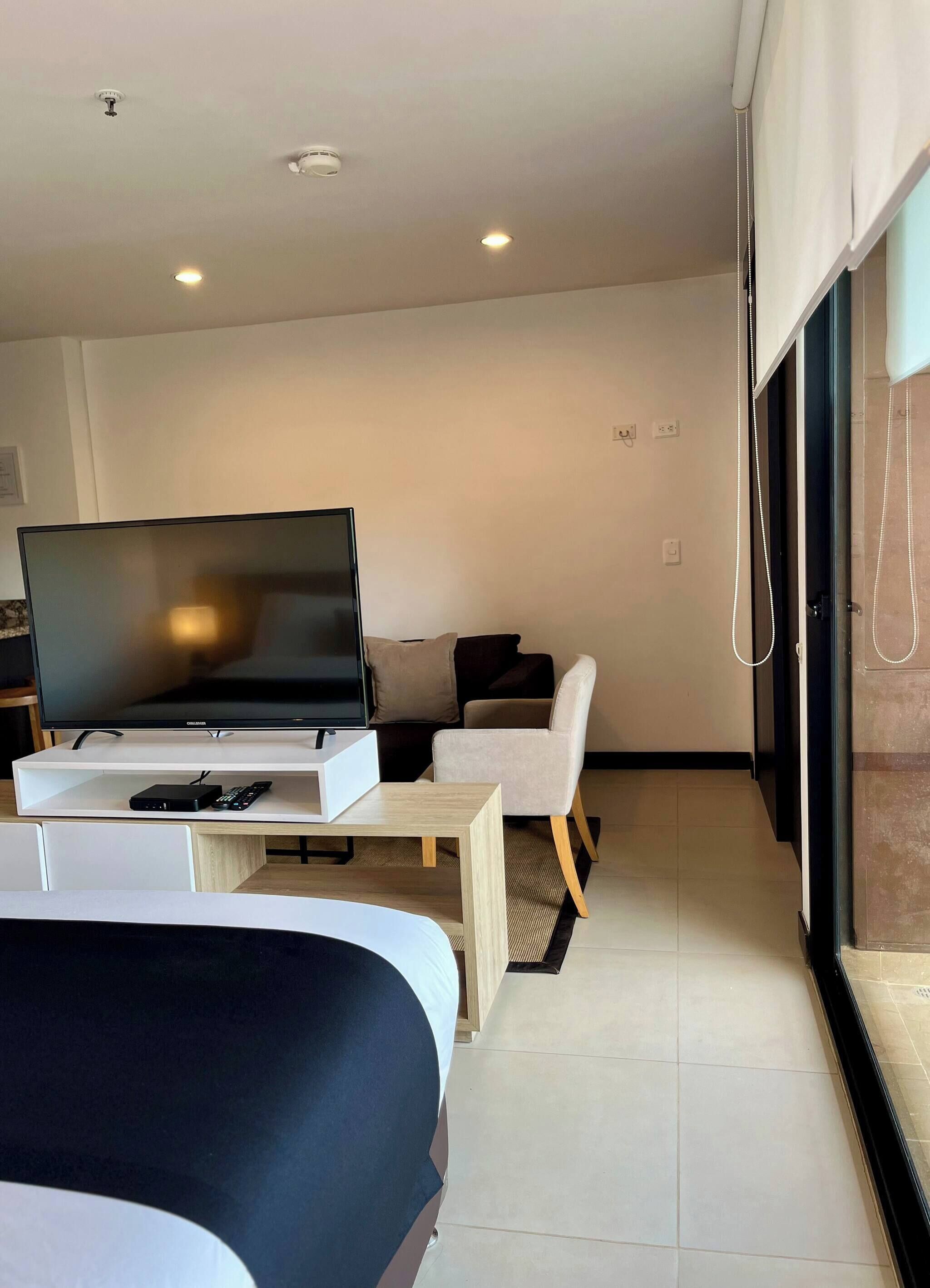 Une chambre d’hôtel moderne, équipée d’un lit, d’un téléviseur posé sur un meuble, d’une petite table, d’une chaise et d’une grande fenêtre.