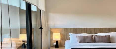 A modern hotel room with a large bed, a bedside table with a lamp, a glass partition, and a view of the outside.