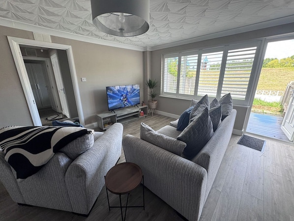 Un salon moderne avec un canapé gris, un téléviseur et une vue sur l’extérieur.