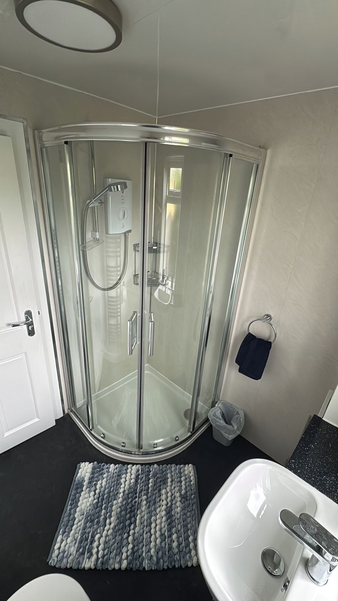Une douche fermée par des parois en verre, dotée d’un pommeau de douche à main, un lavabo blanc avec un robinet chromé et un petit tapis sur le sol.