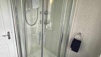 Une douche fermée par des parois en verre, dotée d’un pommeau de douche à main, un lavabo blanc avec un robinet chromé et un petit tapis sur le sol.