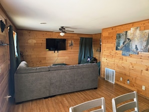 A cozy room with a sectional sofa, a ceiling fan, a flat-screen TV mounted on the wall, and a large window with curtains.
