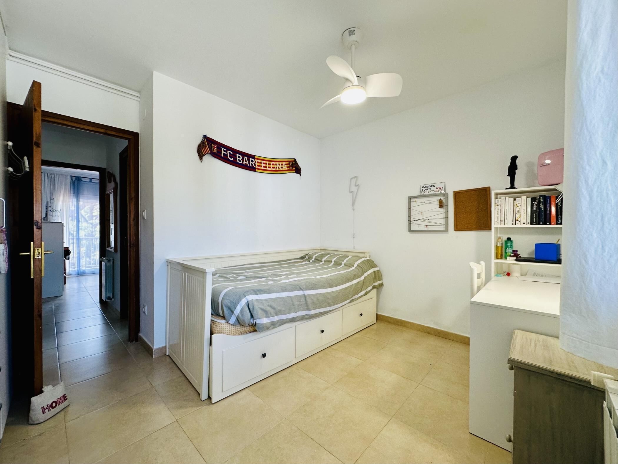 A bedroom with a bed, a ceiling fan, a radiator, a bookshelf, and a door leading to another room.