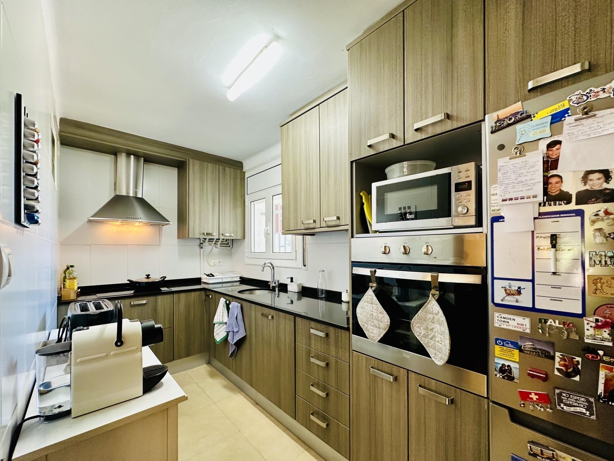 A modern kitchen with wooden cabinets, a microwave, an oven, and a refrigerator with magnets.