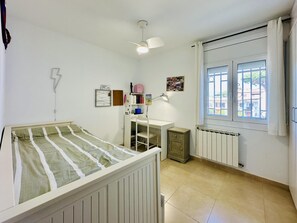Une pièce comprenant un lit, un bureau, une chaise, un radiateur, une fenêtre et un ventilateur de plafond.