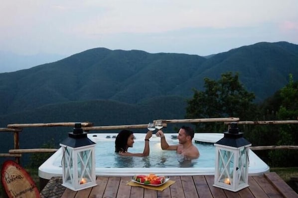 A couple in a hot tub with a mountain view, surrounded by wooden deck and lanterns.