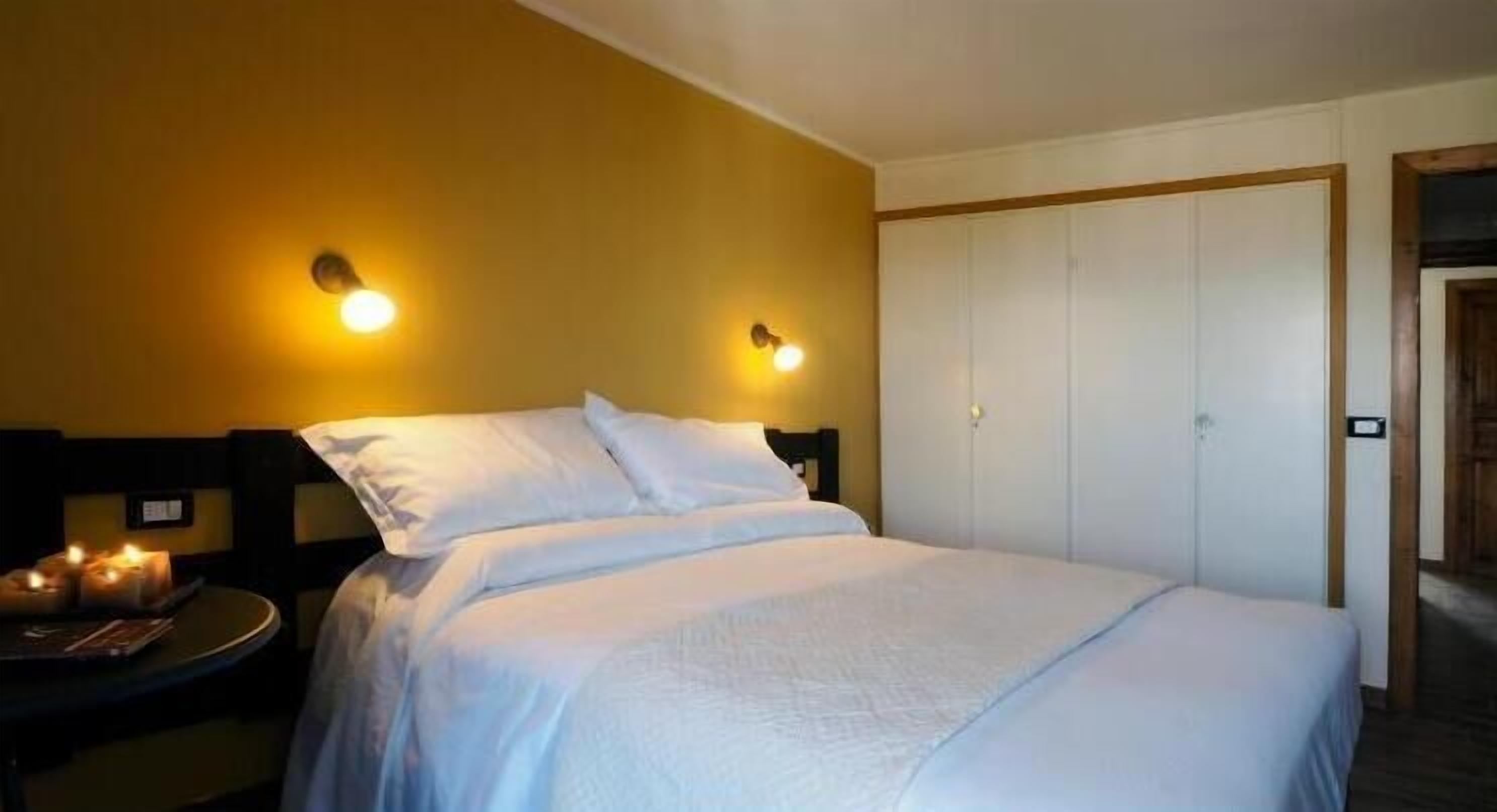 Habitación de hotel con cama, dos lámparas de pared, una mesita de noche con velas y una puerta cerrada.